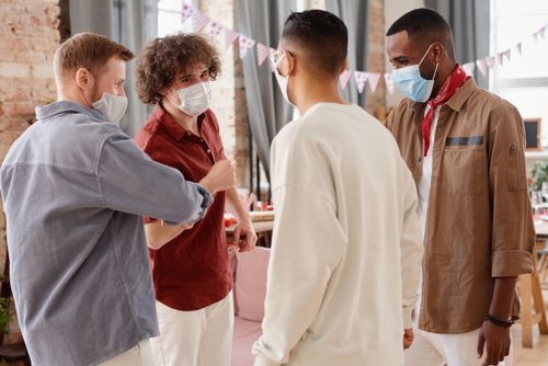 Free Four diverse men wearing masks socializing inside, emphasizing safe gatherings. Stock Photo