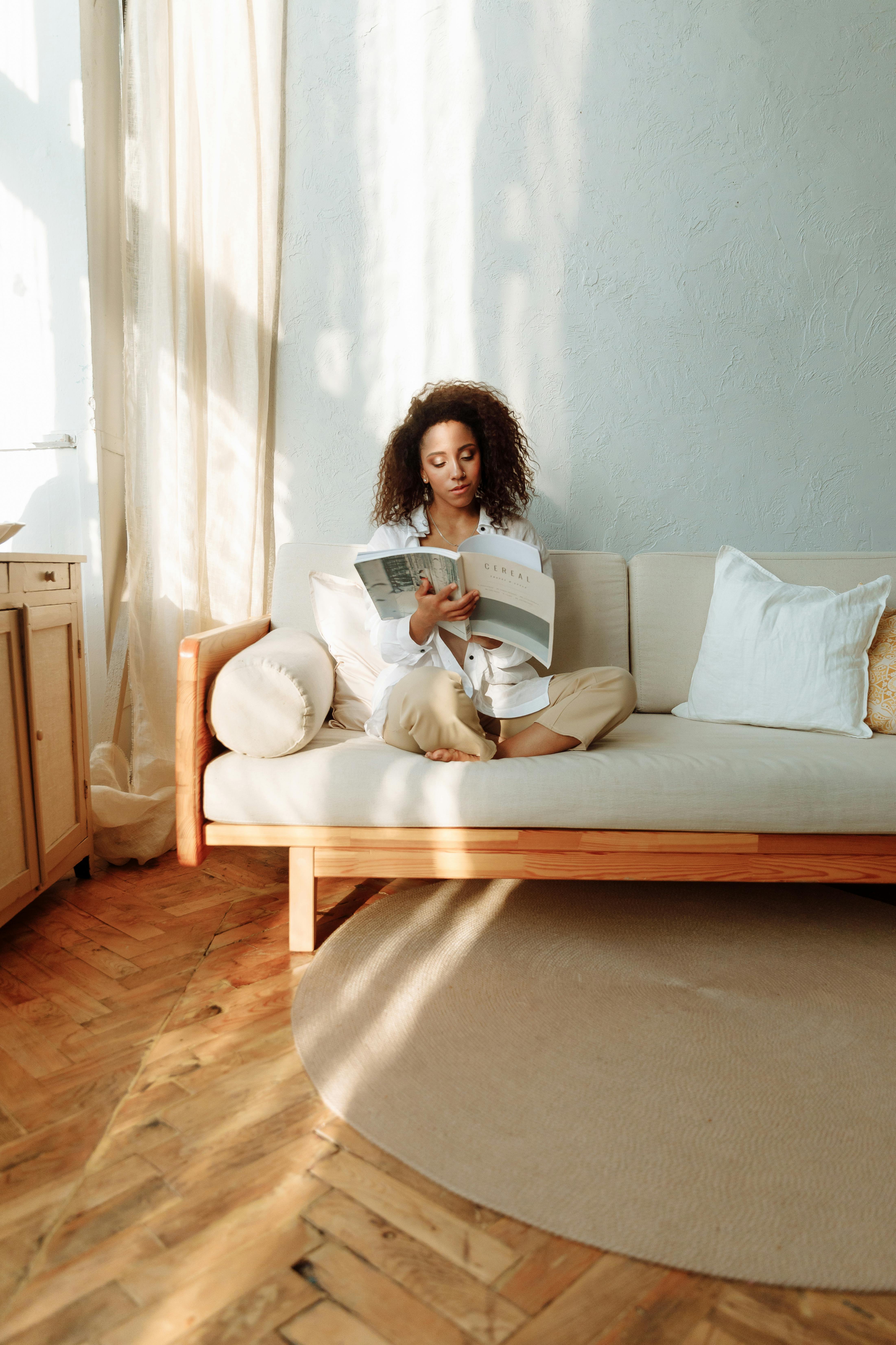 A woman relaxes on a comfortable sofa, enjoying a magazine in a serene, sunlit room.