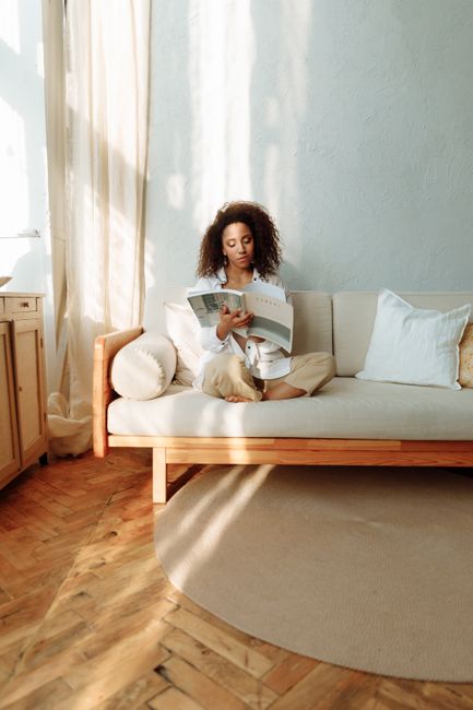 A woman relaxes on a comfortable sofa, enjoying a magazine in a serene, sunlit room.