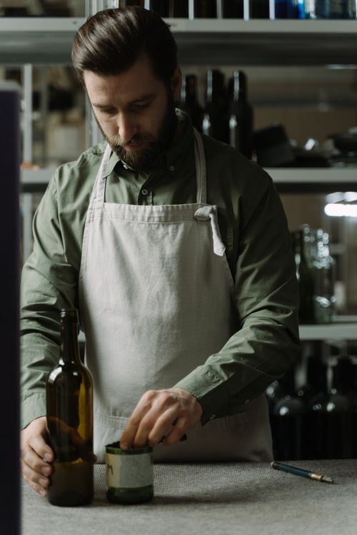 Free A man in an apron arranging bottles inside a room with shelves in the background. Stock Photo