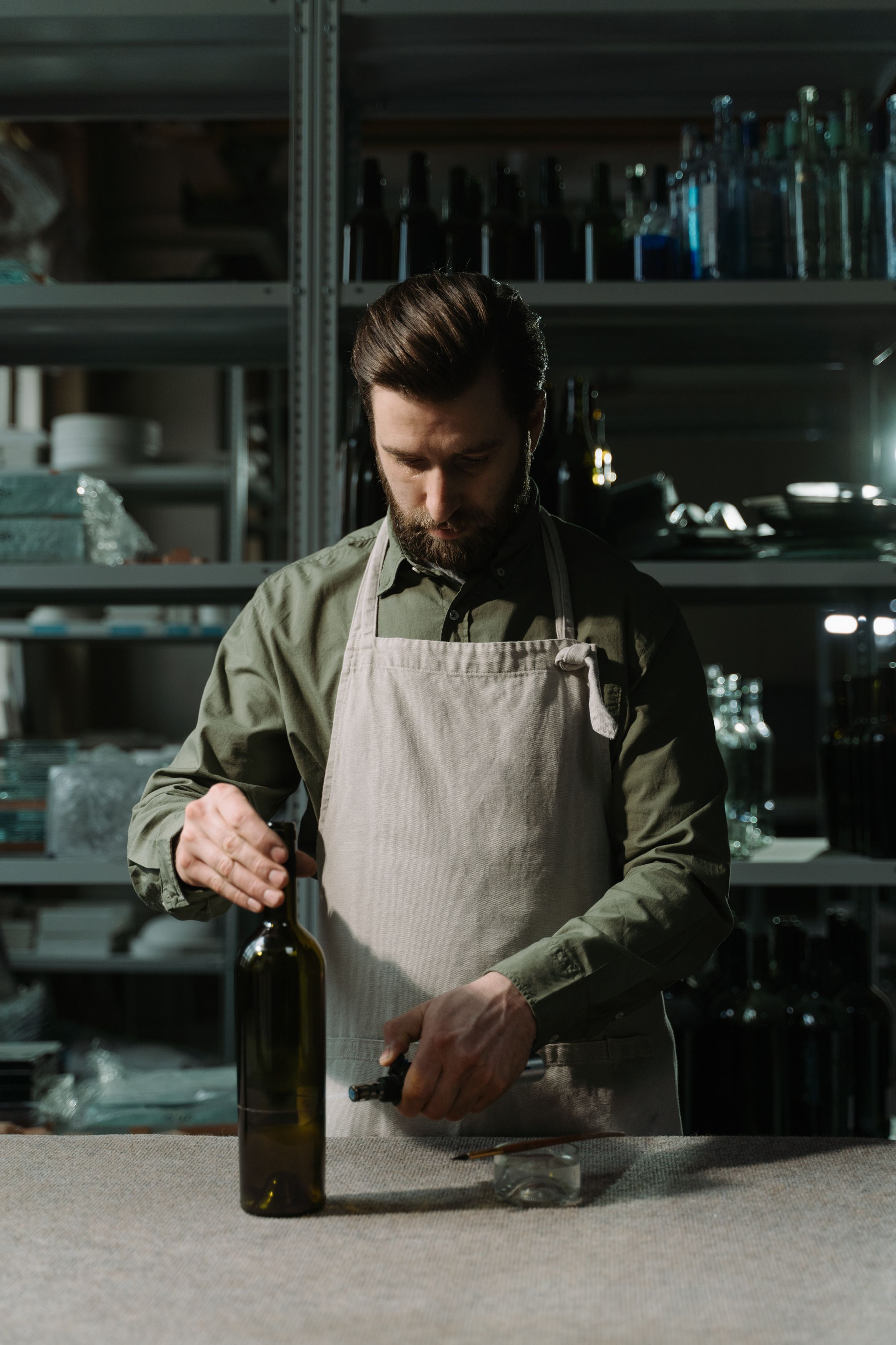 A man in an apron holding a wine bottle inside a vintage-style workshop or kitchen.