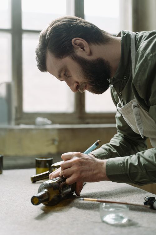 Free Bearded artisan using a glass cutter on a wine bottle in workshop. Stock Photo