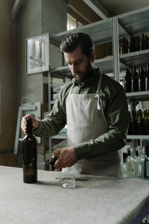 Free Man in apron meticulously crafts bottles in rustic factory setting. Industrial shelves in background. Stock Photo