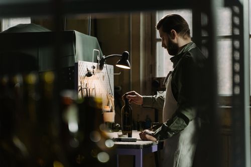 Free Man in apron inspects a glass bottle under workshop lighting, creating a focused industrial atmosphere. Stock Photo
