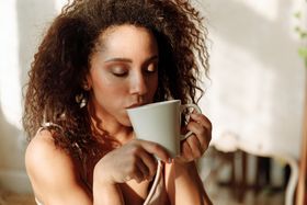 African American woman with curly hair enjoying a hot drink indoors, eyes closed, in warm sunlight.