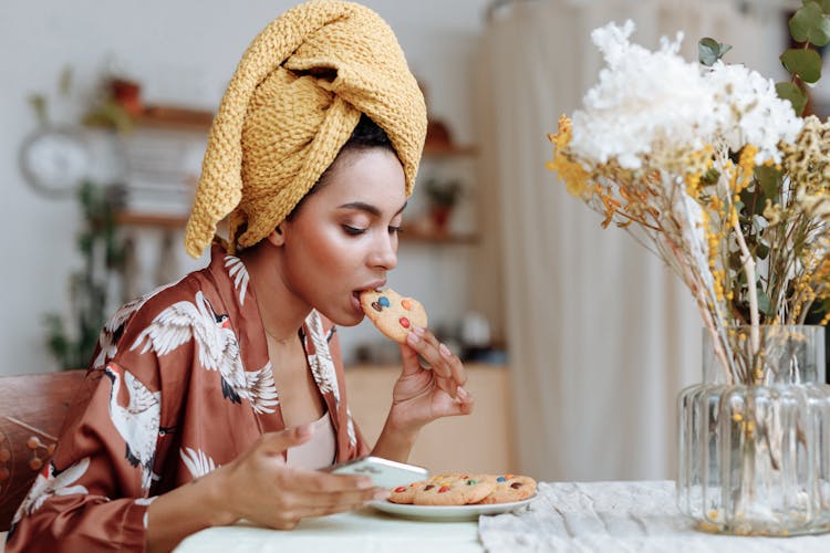 A Woman Eating A Cookie While Using Her Phone