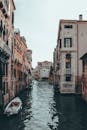 Boats on canal with ripples between old multistory stone building facades under light sky in Venice