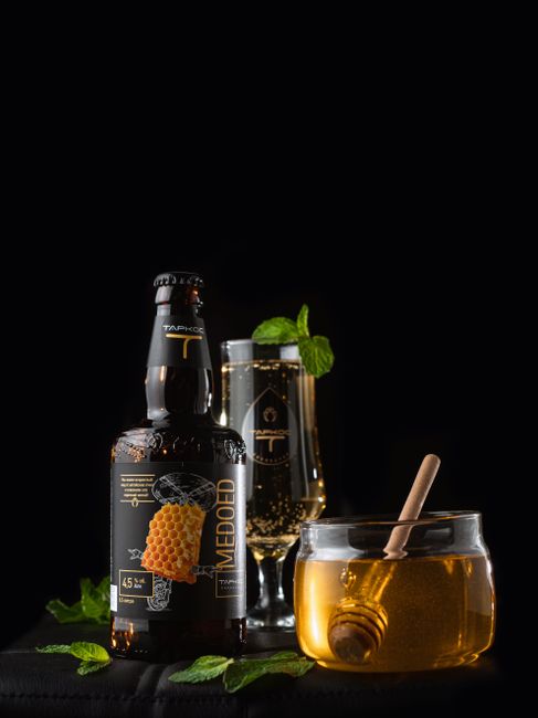 A goblet of amber mead next to a broken honeycomb and a wooden honey dipper on a dark wood table.