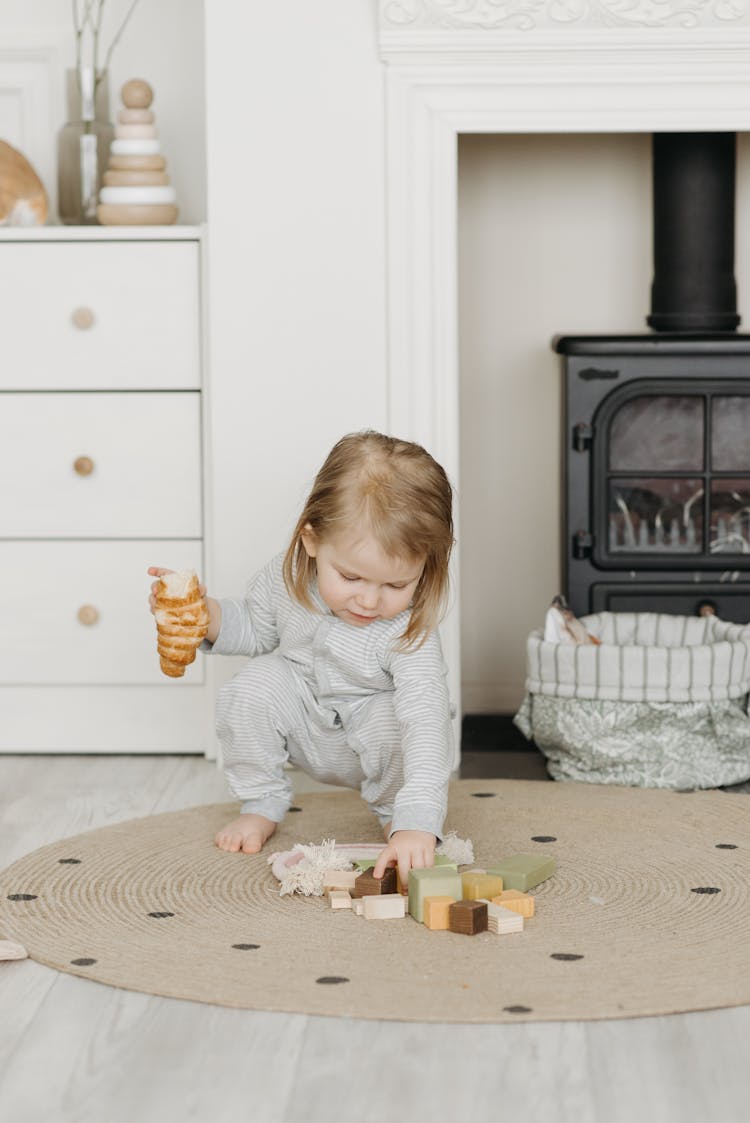 Photo Of A Kid Picking Up Her Toys While Holding A Piece Of Bread