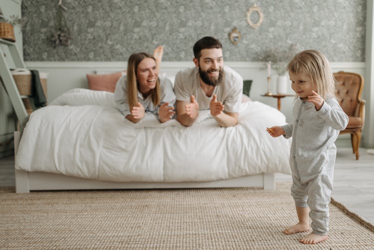 A Couple Lying On The Bed While Looking At Their Daughter Standing