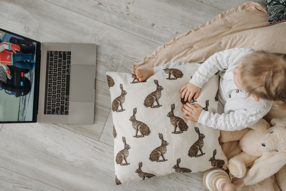A child in a white sweater watching a laptop cartoon, surrounded by toys.