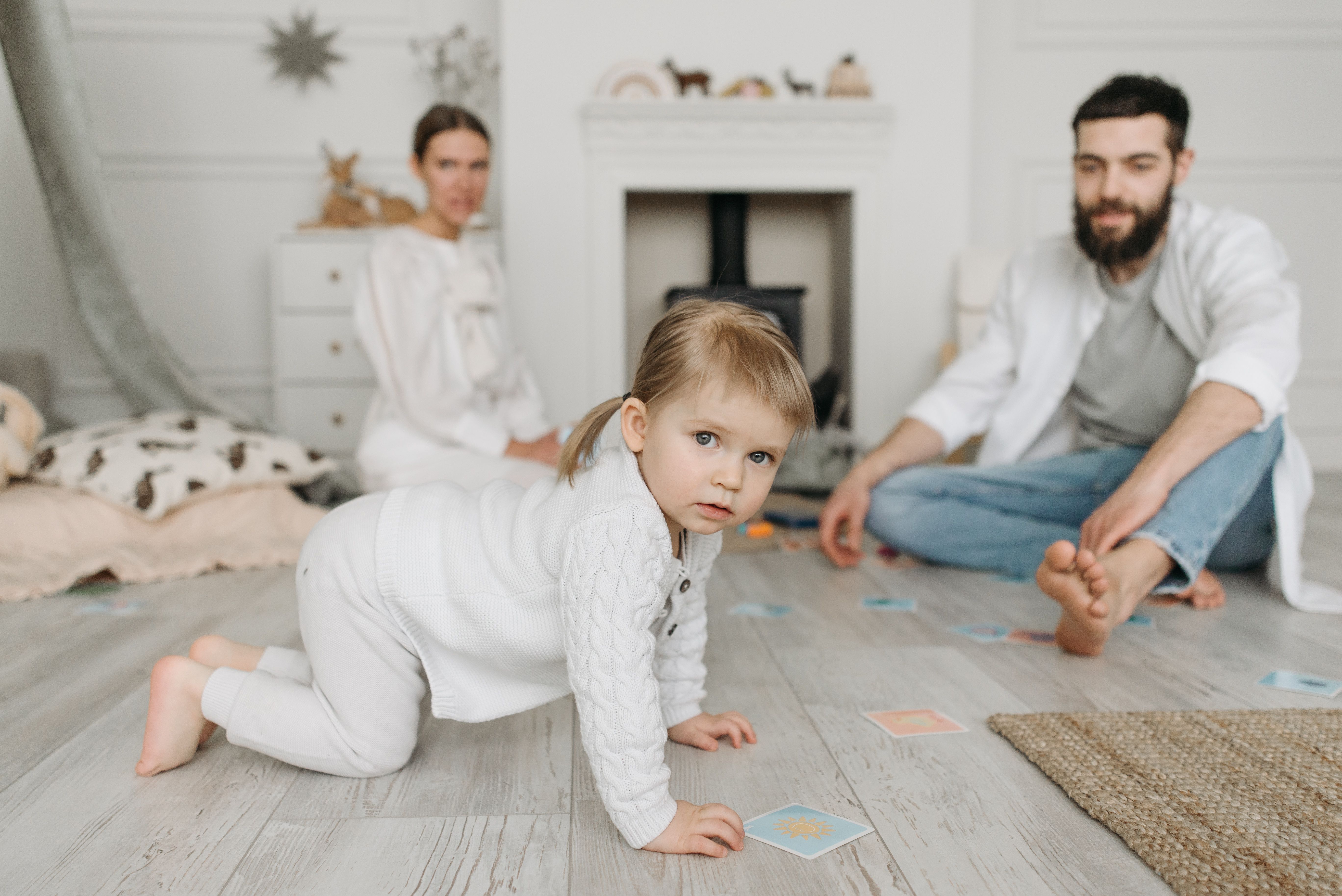Cute toddler in white sweater crawls on the floor, with parents in a cozy room.