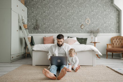 A bearded father works on a laptop while sitting on the bedroom floor with his young daughter, embracing a remote work lifestyle.