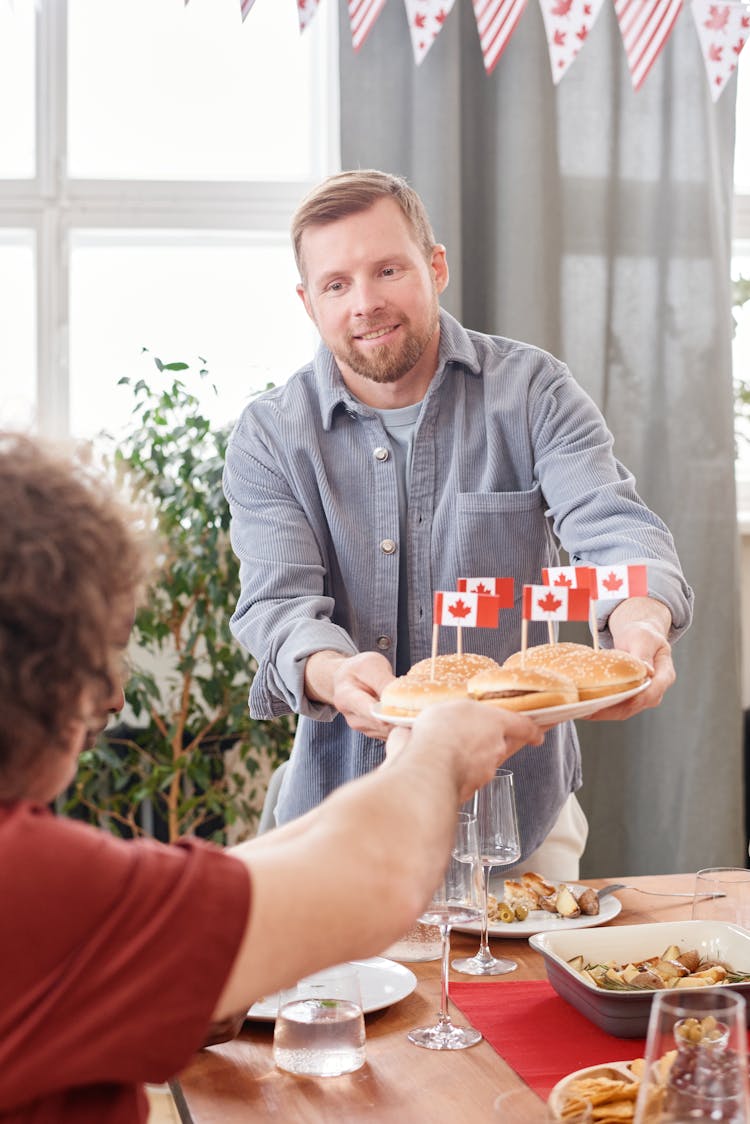 A Bearded Man Passing A Plate Full Of Hamburgers