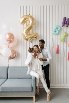 An interracial family celebrating a toddler's second birthday with balloons and decorations.