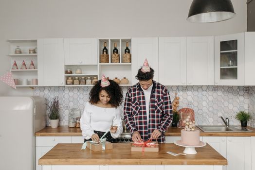 A couple in party hats wrap presents on a kitchen island during a celebration.