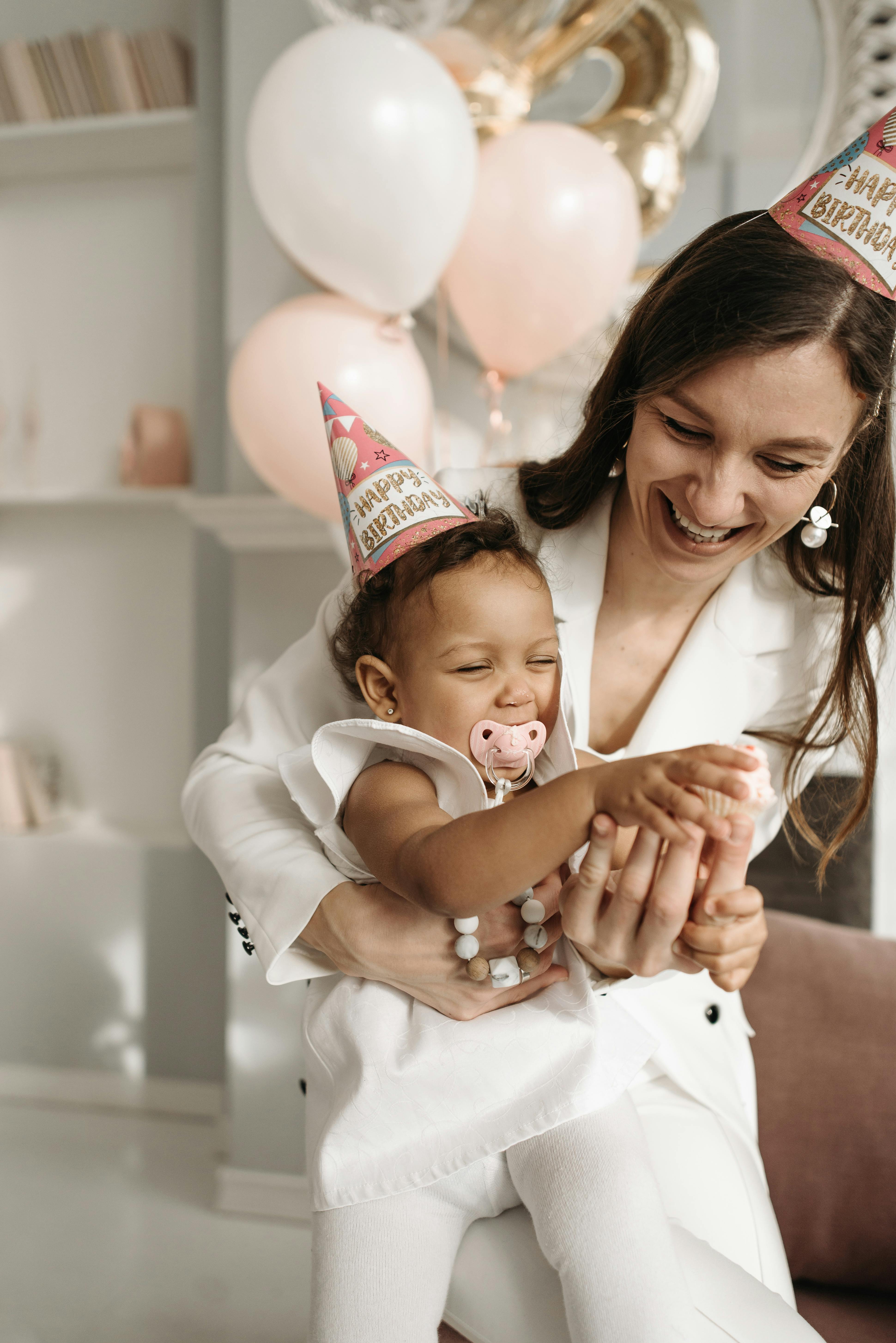 Smiling mother and baby in party hats celebrating a birthday indoors with balloons.
