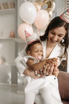 Smiling mother and baby in party hats celebrating a birthday indoors with balloons.