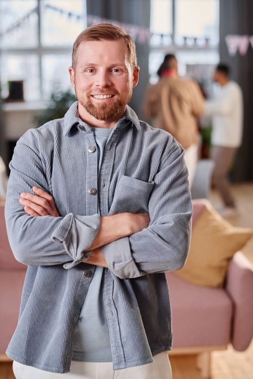 Free Portrait of a smiling man with crossed arms in a casual indoor setting with people in background. Stock Photo