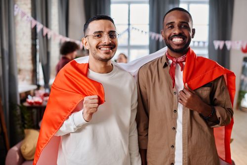 Free Two men smiling, wrapped in flags at a festive indoor event, showcasing diversity and friendship. Stock Photo