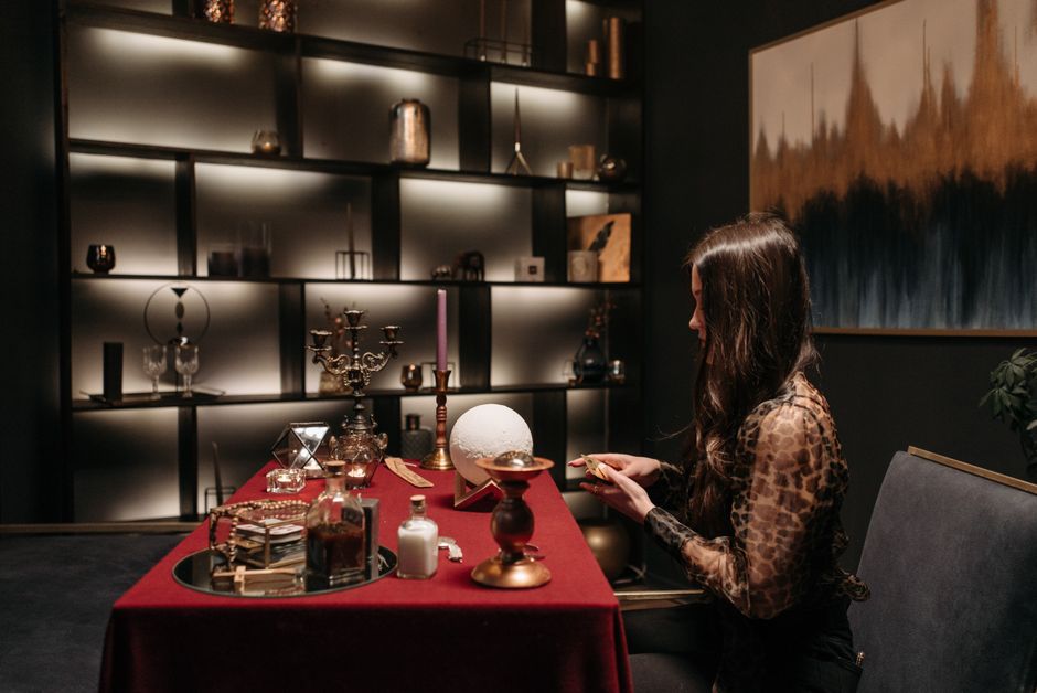 A young woman sitting at a table with tarot cards spread before her, chin resting on her hand in contemplation, expression calm and focused, sunlight streaming through the window