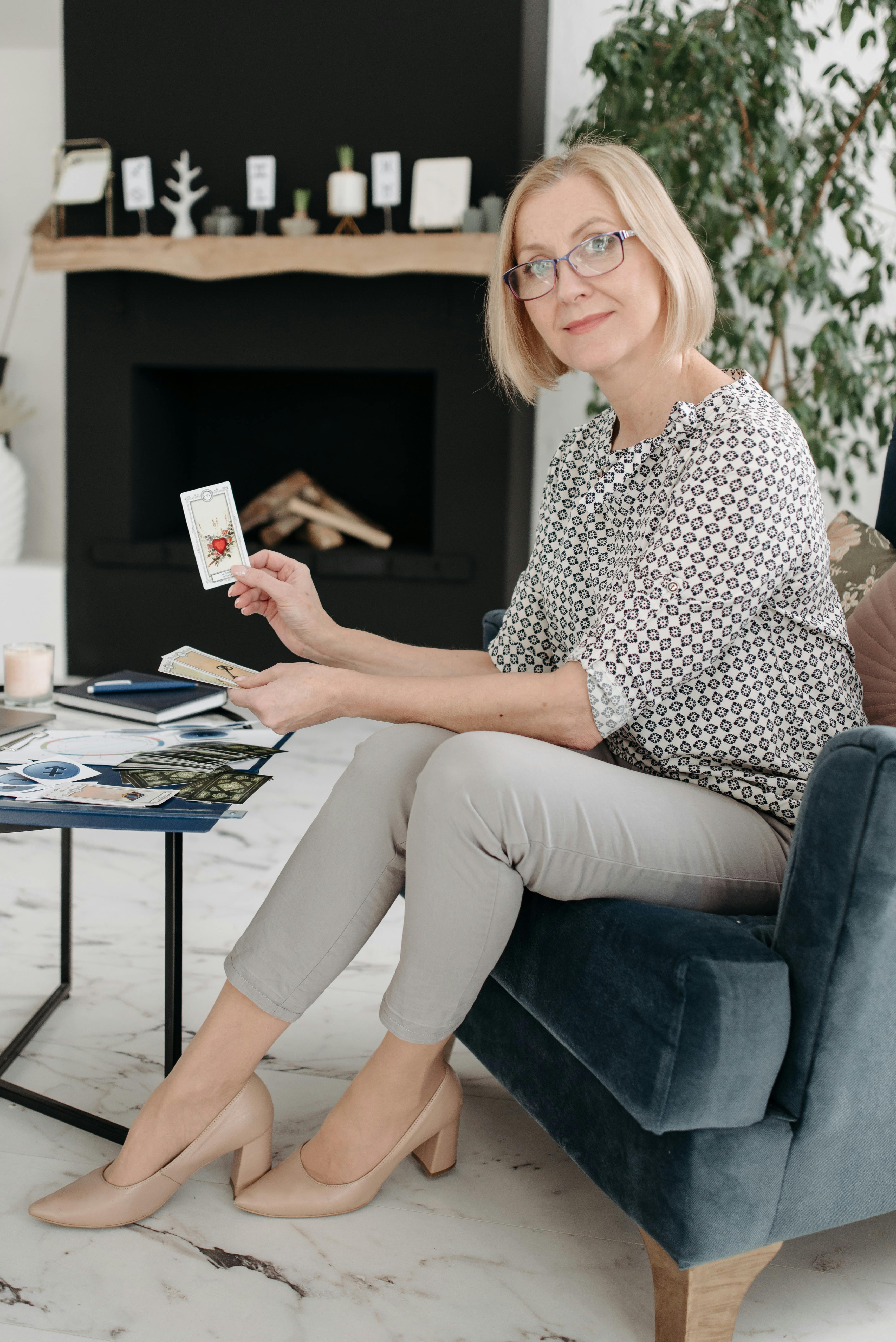 A woman seated in a cozy room, holding tarot cards, suggesting divination or fortune-telling.
