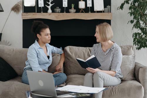 Free Two women sitting on a sofa having a thoughtful discussion, one holding a book. Stock Photo