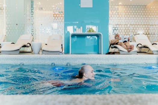 Elderly woman swimming in a modern indoor pool while another senior relaxes on a lounge chair.