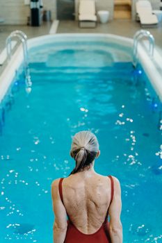 Back view of a woman in a red swimsuit by an indoor swimming pool, ready to dive.