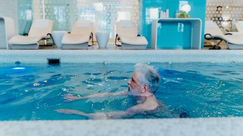 Older man enjoying a swim in a stylish indoor pool facility with relaxation chairs.