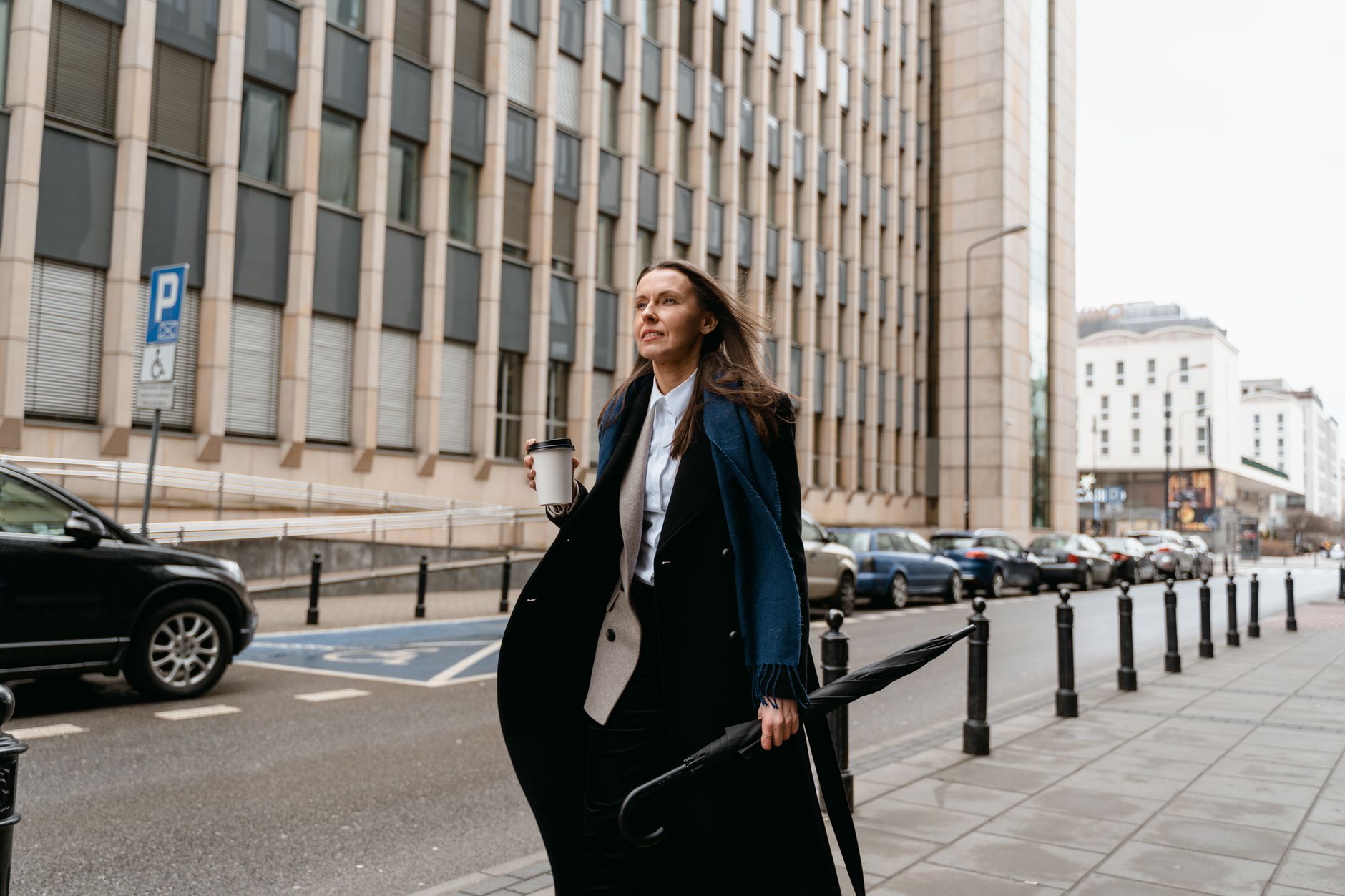 Confident woman walking on city street holding coffee and umbrella.