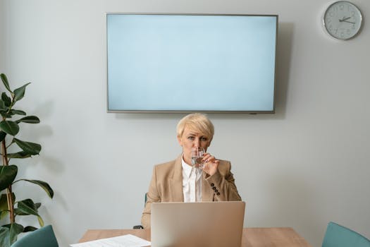 Businesswoman in office setting with laptop, drinking water, with minimalistic decor.