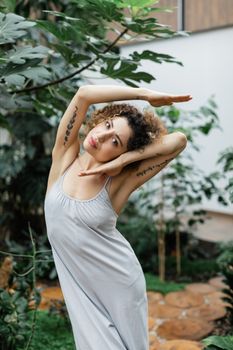 A young woman wearing a flowing dress poses gracefully amidst lush indoor greenery, showcasing elegance and style.