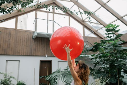 Woman with curly hair exercising with a large red ball in a bright indoor greenhouse setting.