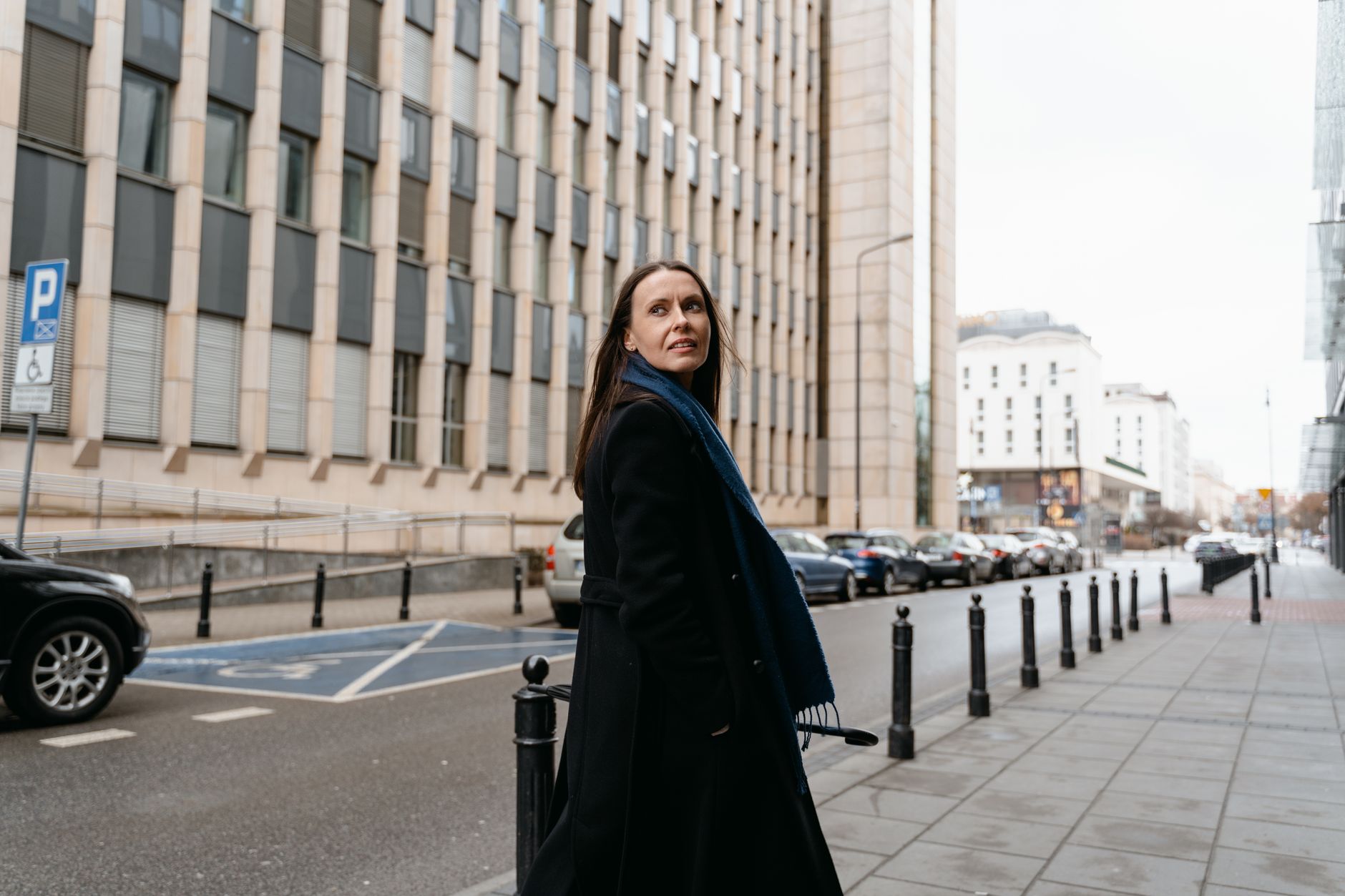 A businesswoman in a black coat and scarf walking down a city street, looking back.