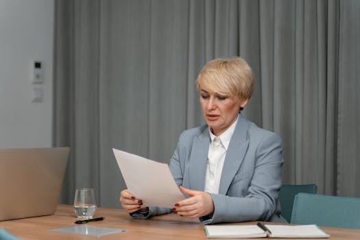 Businesswoman in a blazer reviewing documents at a desk. Professional office setting with laptop and papers.