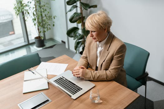 Professional woman in office setting working on a laptop, surrounded by office items.