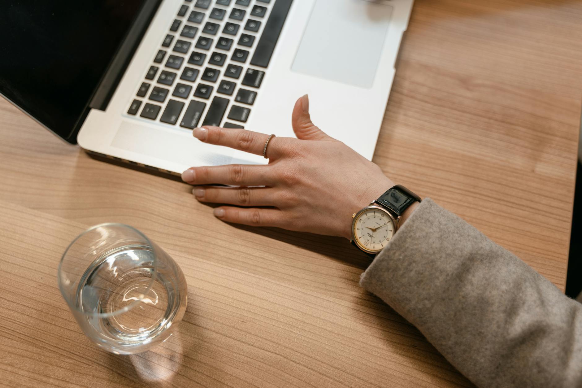 A close-up of a woman's watch-adorned hand on a wooden desk beside a laptop and glass of water.