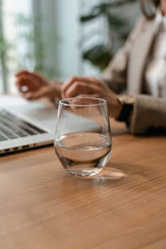 Close-up of a glass of water on a wooden desk with hands typing on a laptop in a modern office setting.