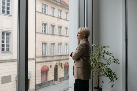 A thoughtful woman stands by a large window watching the cityscape outside, arms crossed