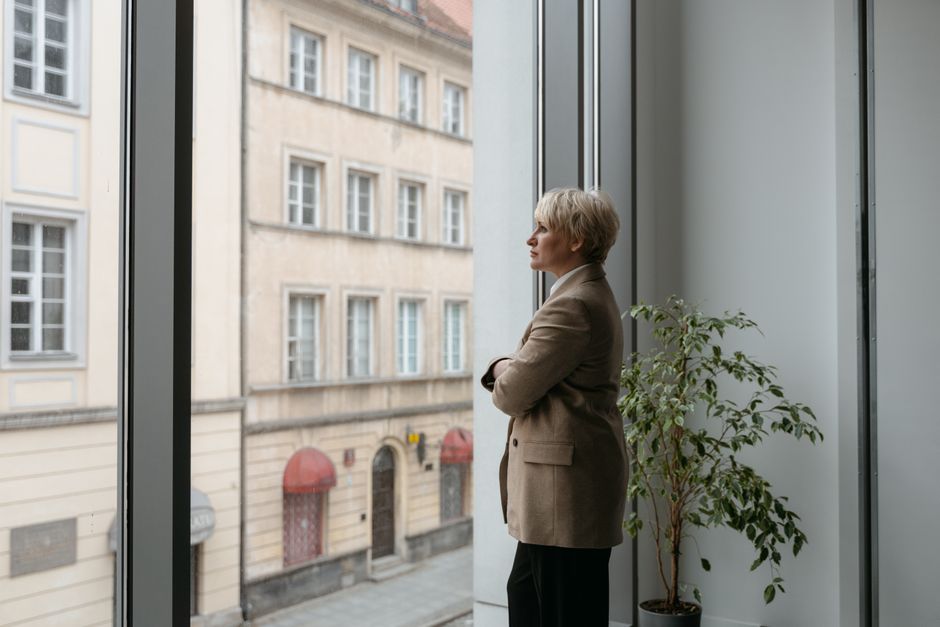 A thoughtful woman stands by a large window watching the cityscape outside, arms crossed