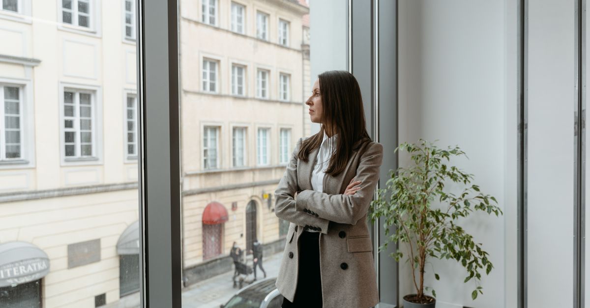 A woman in a blazer stands by a large window, gazing at the cityscape from an office.