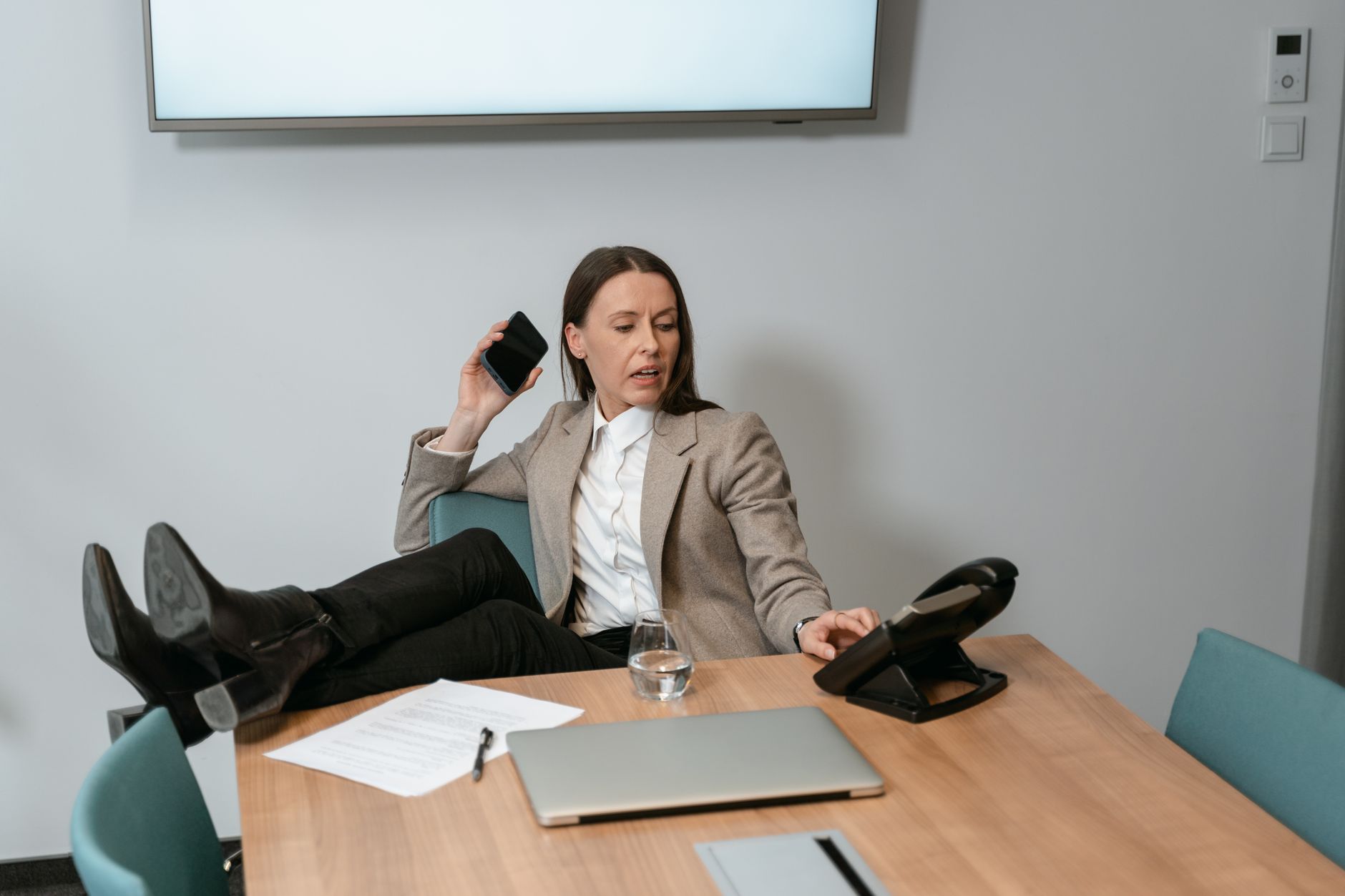 A businesswoman in a blazer multitasks with a smartphone and desk phone in a modern office setting.