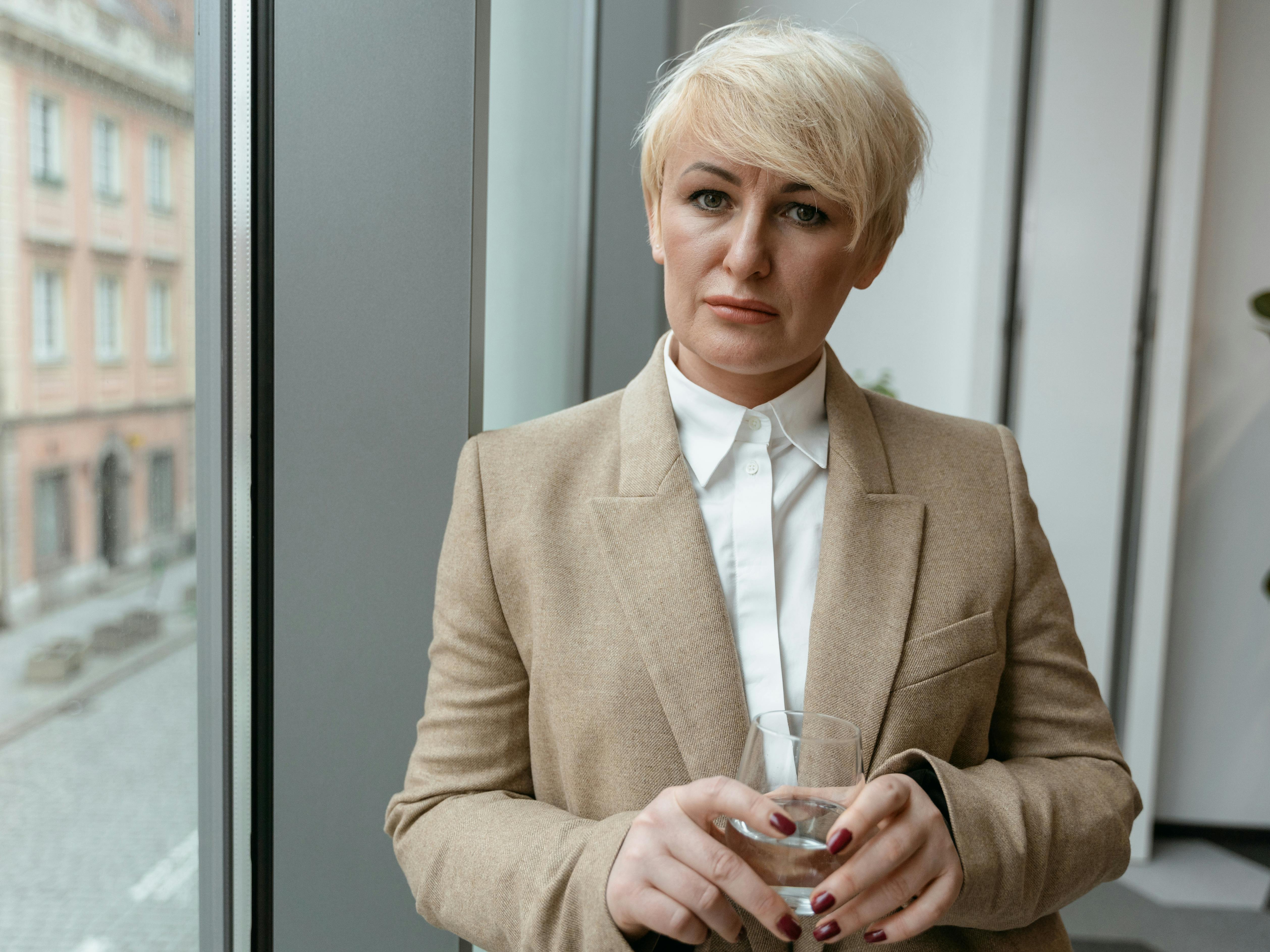 a serious woman in brown blazer holding a glass of water looking at camera