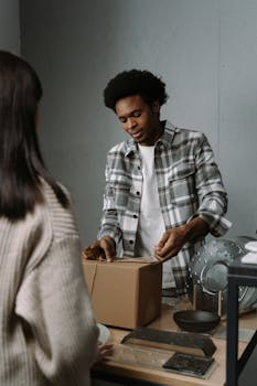 A man in a plaid shirt packs a box in a room, viewed from the side.