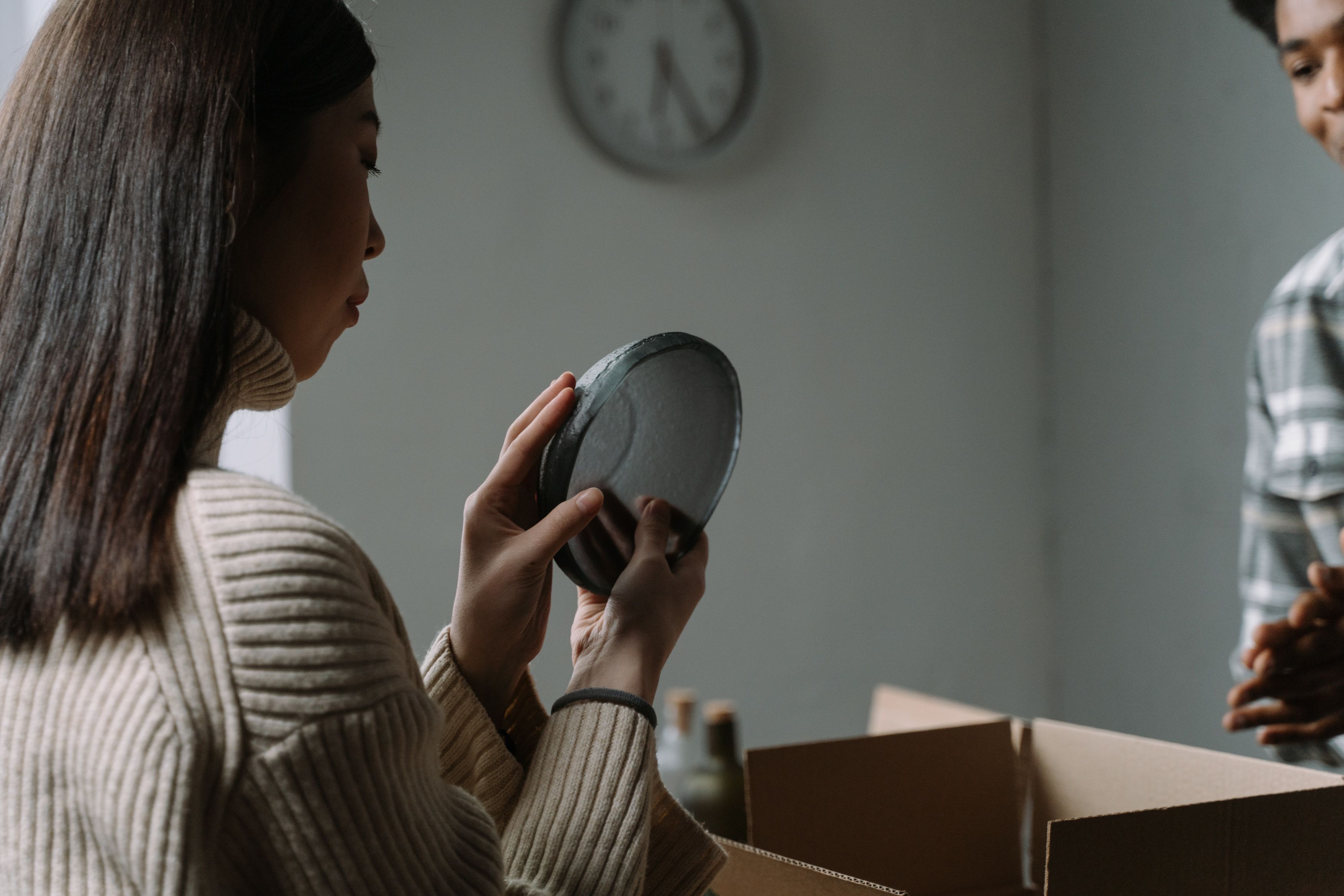 A woman carefully unpacks dishware from a cardboard box in a cozy indoor setting.