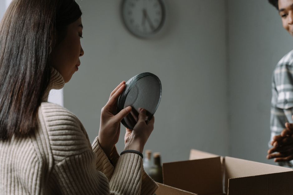 A woman carefully unpacks dishware from a cardboard box in a cozy indoor setting.