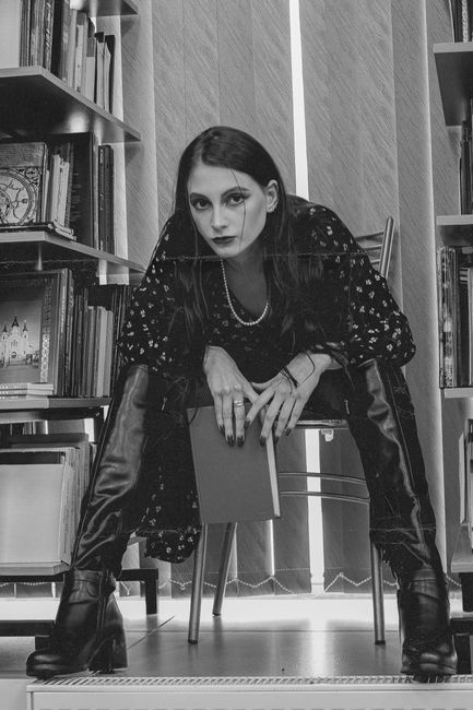 Black and white of confident woman in leather boots with textbook looking at camera indoors