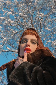 From below of young female with bright makeup in outerwear looking at camera under winter tree in sunlight