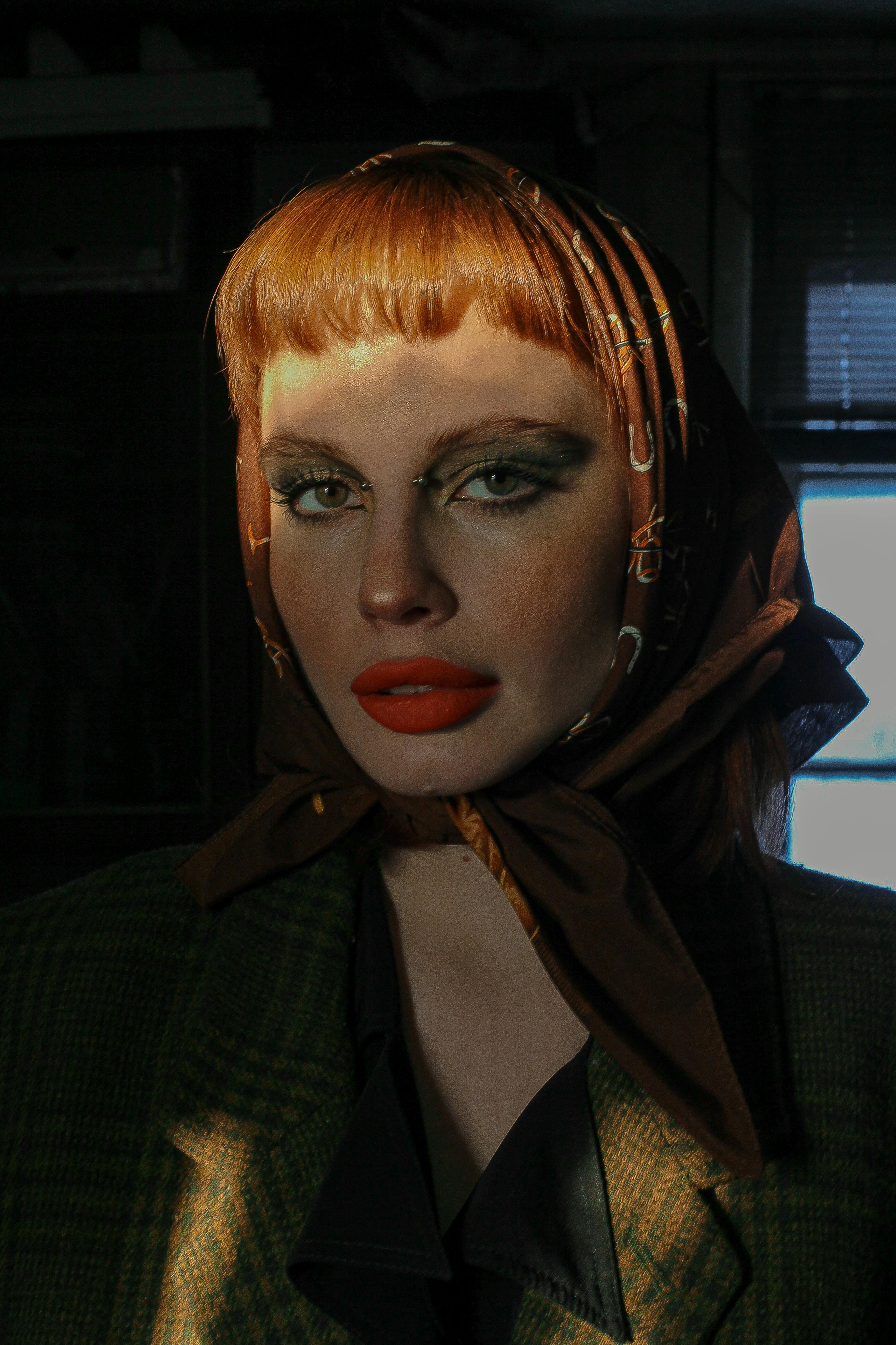 Young trendy woman with red lips and piercings in checkered jacket looking at camera in sun rays indoors
